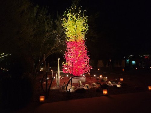 A vibrant, colorful glass sculpture resembling a cactus, illuminated at night with surrounding lights and desert plants.