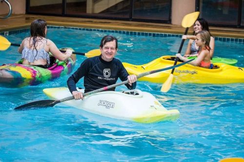 A man in a kayak smiles while others paddle in a pool, enjoying a kayaking activity.