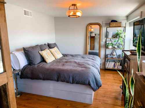 Cozy bedroom featuring a large bed, decorative pillows, a mirror, shelves with plants, and warm lighting.