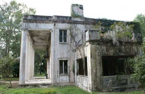 Abandoned building with crumbling columns, overgrown vegetation, and a weathered facade in a lush green setting.