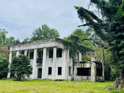 An abandoned, ivy-covered building with columns, surrounded by trees and overgrown grass under a cloudy sky.