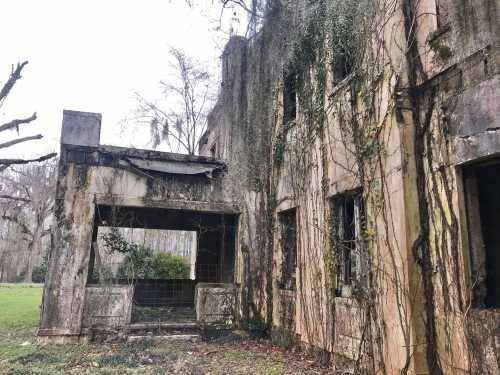 Abandoned building covered in vines, with crumbling walls and a partially collapsed porch, set in a grassy area.