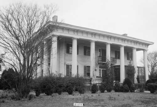 A historic two-story house with large columns, surrounded by trees and shrubs, in black and white.