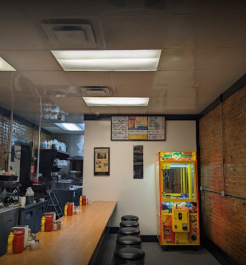 A diner interior with a long wooden counter, stools, condiments, and a colorful claw machine in the corner.