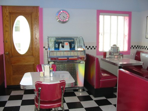 A retro diner interior featuring red booths, a jukebox, and a checkered floor, with a wooden door and wall clock.