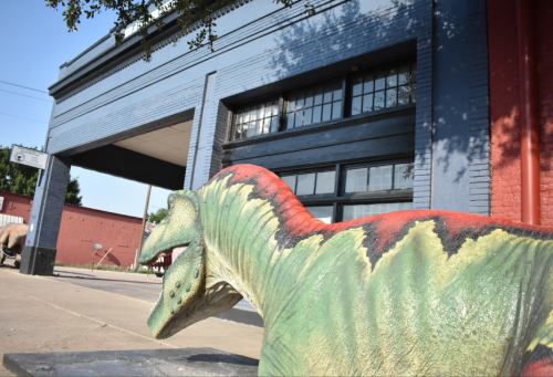 A colorful dinosaur sculpture in front of a blue and red building with large windows.