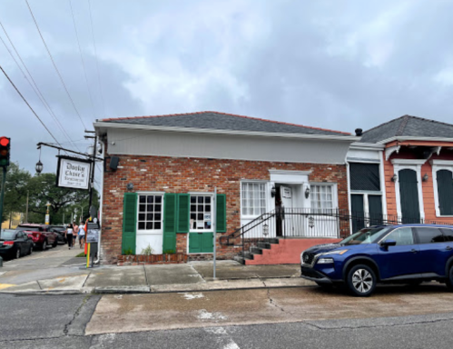 A brick building with green shutters and a sign, located at a street corner with parked cars and cloudy skies.