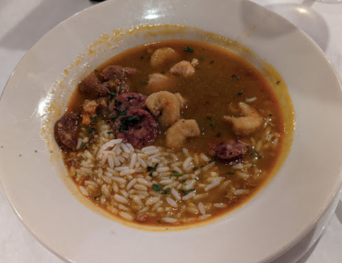 A bowl of gumbo with shrimp, sausage, and rice, garnished with herbs in a rich, flavorful broth.