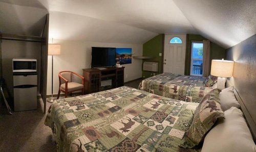 Cozy hotel room with two beds, a TV, mini fridge, and a window, featuring rustic decor and warm lighting.