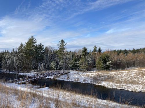 A snowy landscape featuring a wooden bridge over a calm river, surrounded by evergreen trees and a clear blue sky.