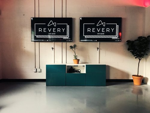 Two screens displaying "REVERY VR BAR" above a modern cabinet with a plant and a potted tree in a minimalist room.