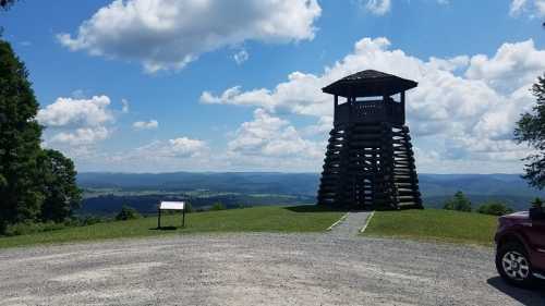 A wooden observation tower on a grassy hill, overlooking a valley with rolling hills and a blue sky filled with clouds.