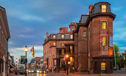 Historic brick building at dusk, featuring flags and street lamps, with cars passing by on a charming street.