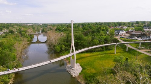 Aerial view of a modern suspension bridge over a river, surrounded by greenery and a small town in the background.