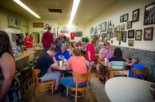 A busy café with patrons seated at tables, chatting, and enjoying drinks, surrounded by framed photos on the walls.