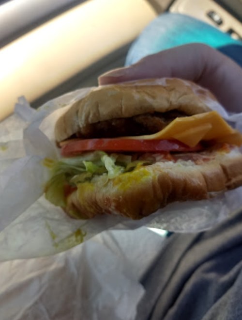 A hand holding a sandwich with lettuce, tomato, cheese, and pickles, wrapped in paper, inside a car.