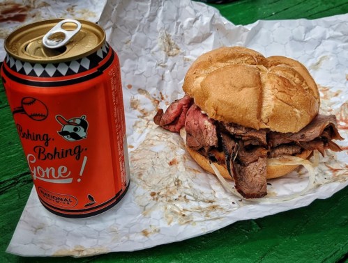 A sandwich with sliced meat on a bun next to a can of soda, all on crumpled paper on a green table.