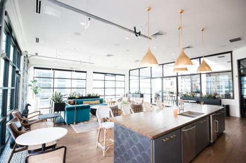 Bright, modern office lounge with large windows, stylish furniture, and a kitchen area featuring pendant lights.