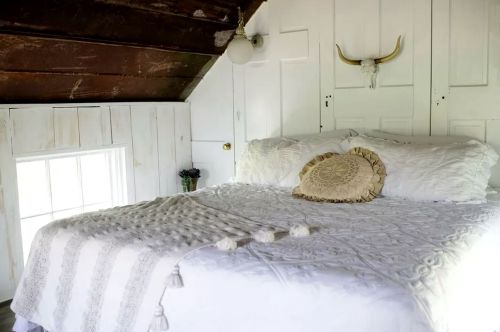 Cozy bedroom with a white bed, decorative pillow, and rustic wooden ceiling. Natural light from a window.