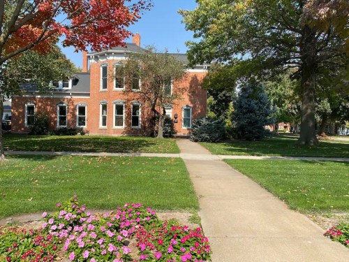 A brick building surrounded by green grass and colorful flowers on a sunny day, with trees in the background.