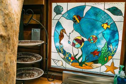 Stained glass window featuring colorful fish and underwater plants, with decorative plates on a shelf nearby.
