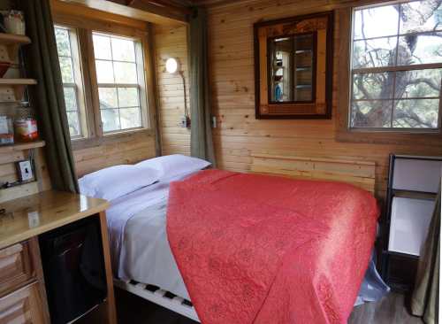Cozy wooden interior of a small room featuring a bed with a red blanket, windows, and a mirror.