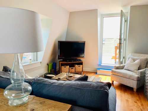 Cozy living room with a gray sofa, lamp, TV, and a striped armchair by a window. Bright and inviting atmosphere.