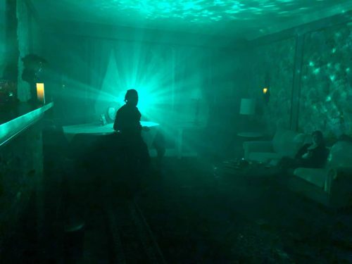 A dimly lit room with green light beams, featuring a person sitting at a table and another figure on a couch.
