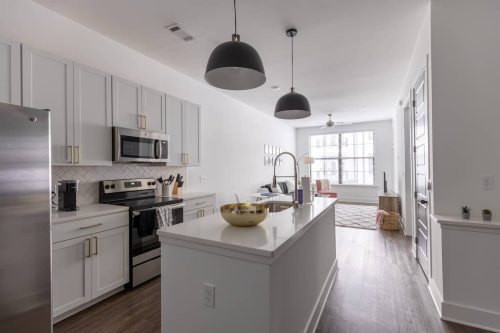 Modern kitchen with stainless steel appliances, white cabinets, and a spacious layout, leading to a bright living area.