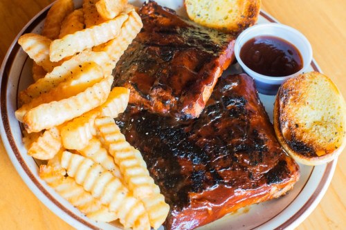 A plate of BBQ ribs with a side of crinkle-cut fries and toasted bread, accompanied by a small cup of barbecue sauce.