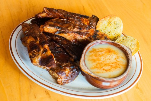 A plate of grilled ribs with a side of creamy sauce and two pieces of cornbread on a wooden table.