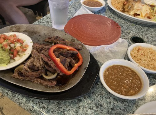 A sizzling platter of grilled meat with red peppers, served with guacamole, tomatoes, rice, and beans on a table.