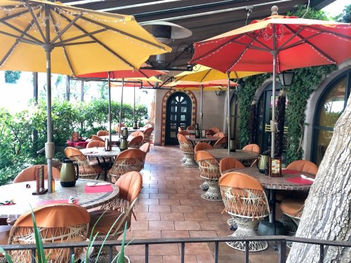 Outdoor restaurant patio with colorful umbrellas, wicker chairs, and tables surrounded by greenery.