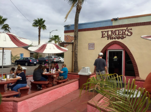 A colorful taco restaurant with outdoor seating, palm trees, and customers enjoying their meals.