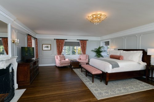 Spacious hotel room with a king bed, two armchairs, a TV, and elegant decor, featuring a chandelier and hardwood floors.