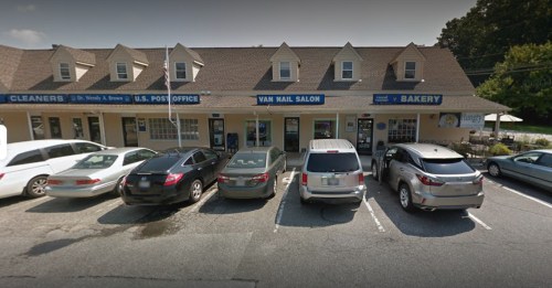 A row of shops including a bakery and a nail salon, with parked cars in front and a clear blue sky above.