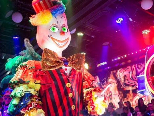 A colorful clown figure with a big smile and vibrant costume, set against a lively, illuminated background.