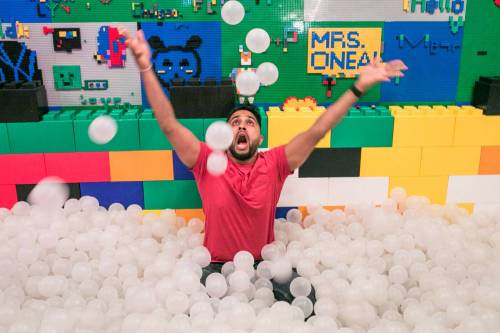 A man in a pink shirt joyfully throws white balls in a colorful ball pit surrounded by LEGO walls.
