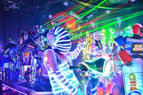 A vibrant scene with performers in glowing costumes and robots, surrounded by colorful laser lights and neon effects.