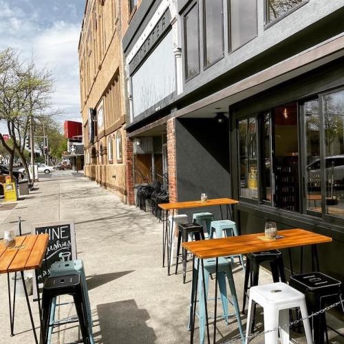 Outdoor seating area with wooden tables and stools beside a brick building on a sunny street.