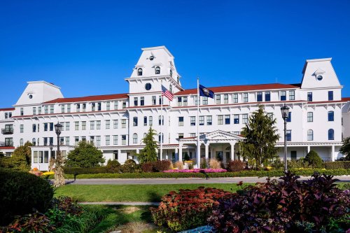A large, white hotel building with a red roof, surrounded by gardens and blue sky. Flags are displayed in front.