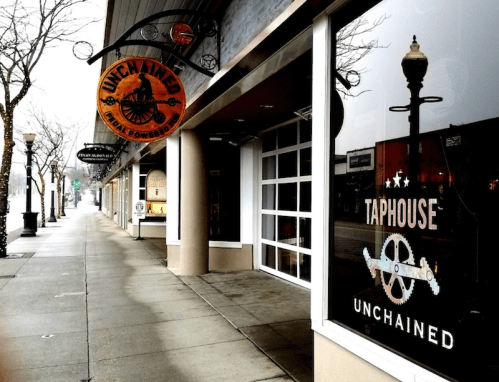 Exterior of Unchained Taphouse, featuring a sign and large windows, on a quiet street with bare trees.