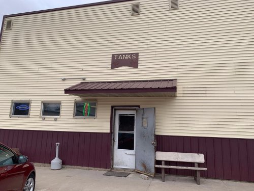 Exterior of a building with a sign reading "TANKS," featuring a door, windows, and a bench outside.