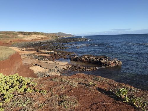 Coastal landscape featuring rocky shorelines, clear blue water, and green vegetation under a clear sky.