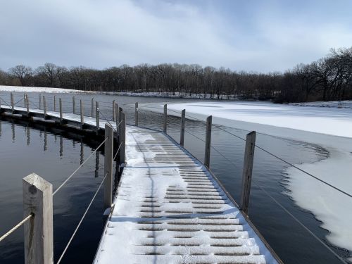 A snowy wooden walkway extends over a calm lake, with ice along the shore and bare trees in the background.
