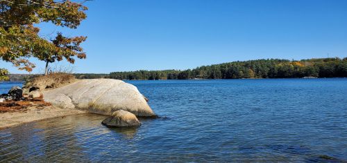 A serene lake scene with a large rock in the foreground, surrounded by trees and clear blue skies.