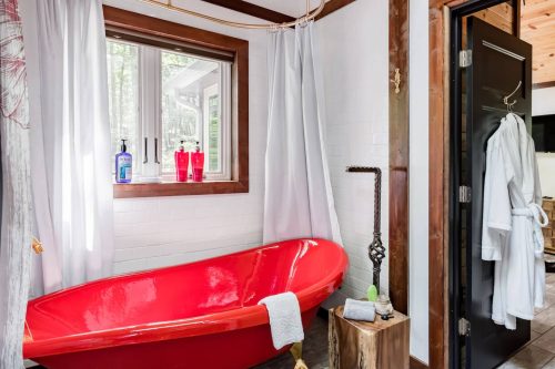 A bright red bathtub in a modern bathroom, with white curtains, a window, and a robe hanging on the door.