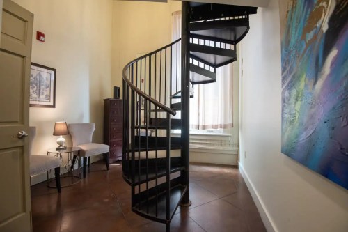 A spiral staircase in a well-lit room with a chair, lamp, and colorful wall art.