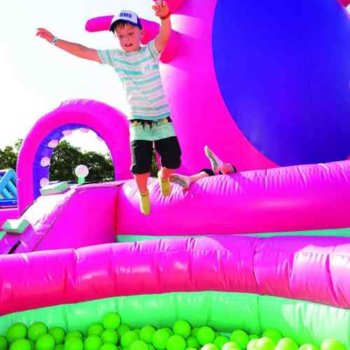A child jumps joyfully from a colorful inflatable structure into a pit filled with green balls.