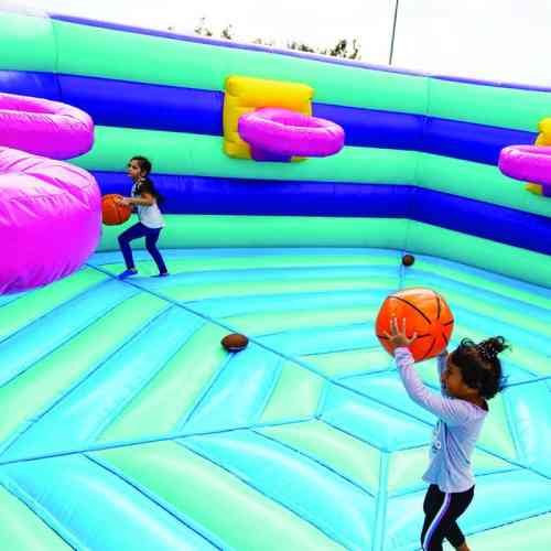 Two children play inside a colorful inflatable bounce house, holding basketballs and preparing to shoot.